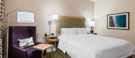 Una habitación de hotel con una cama grande, una silla con un cojín, una mesita de noche con una lámpara y un cuadro en la pared.