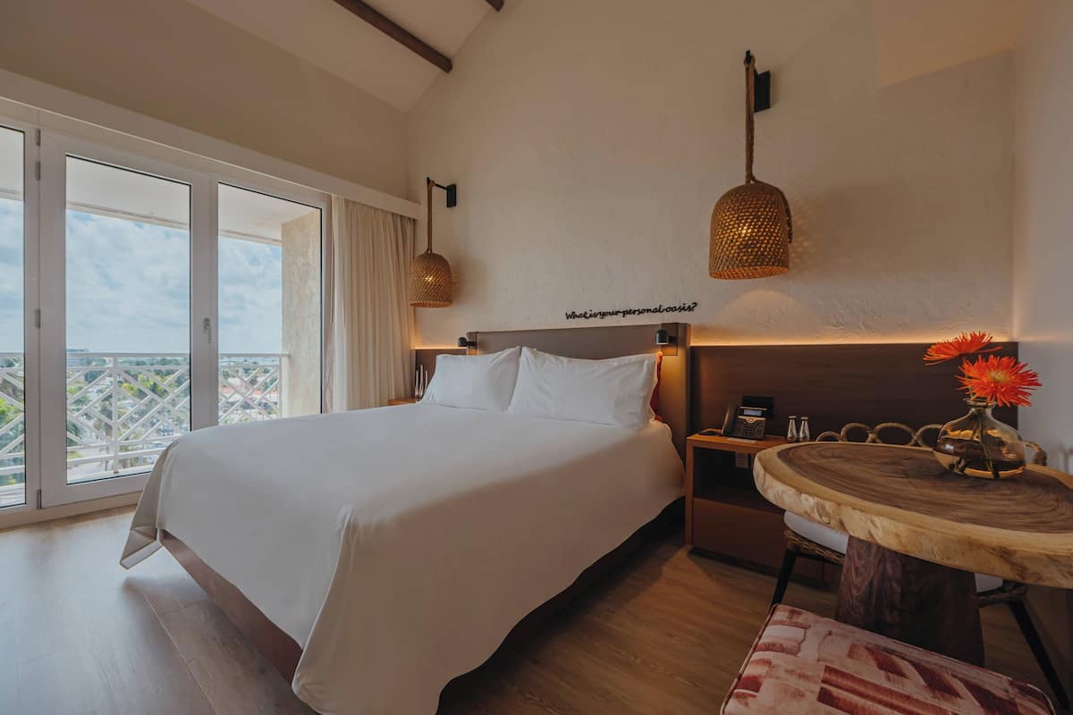 A bedroom with a large bed, a round wooden table with a vase, and a view of the city through the window.