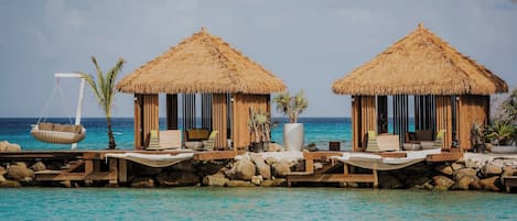 Deux huttes à toit de chaume, dotées de balustrades en bois, chacune comprenant une chaise longue et un fauteuil suspendu, et situées sur une terrasse en bois au-dessus de l’eau.