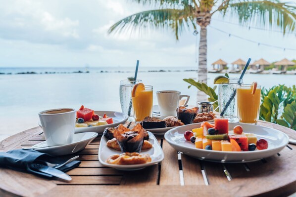 A breakfast table with various foods and drinks, overlooking a beach with umbrellas and palm trees.