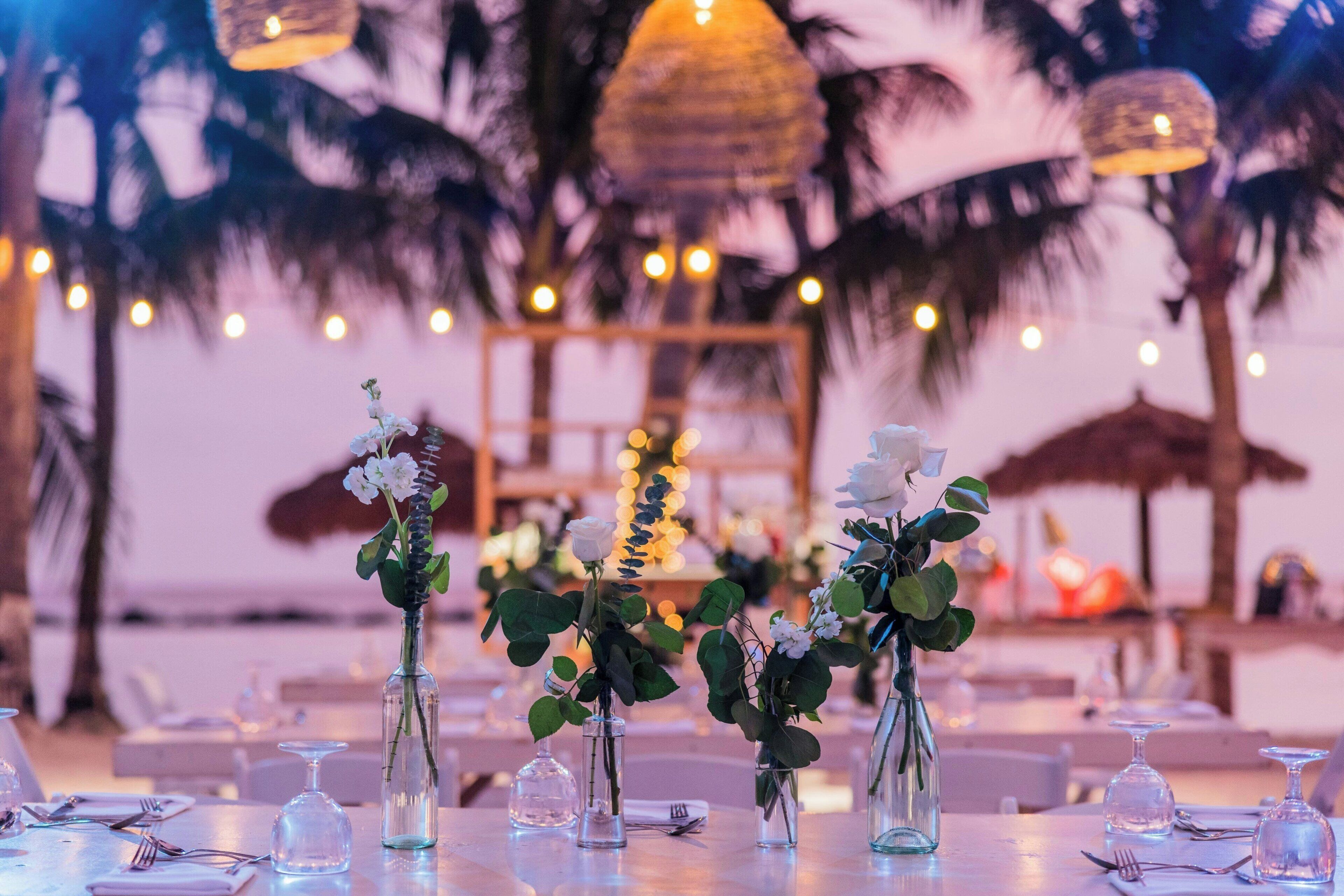 A table set for dining with clear glass vases containing flowers and greenery, and a tropical backdrop with palm trees and string lights.