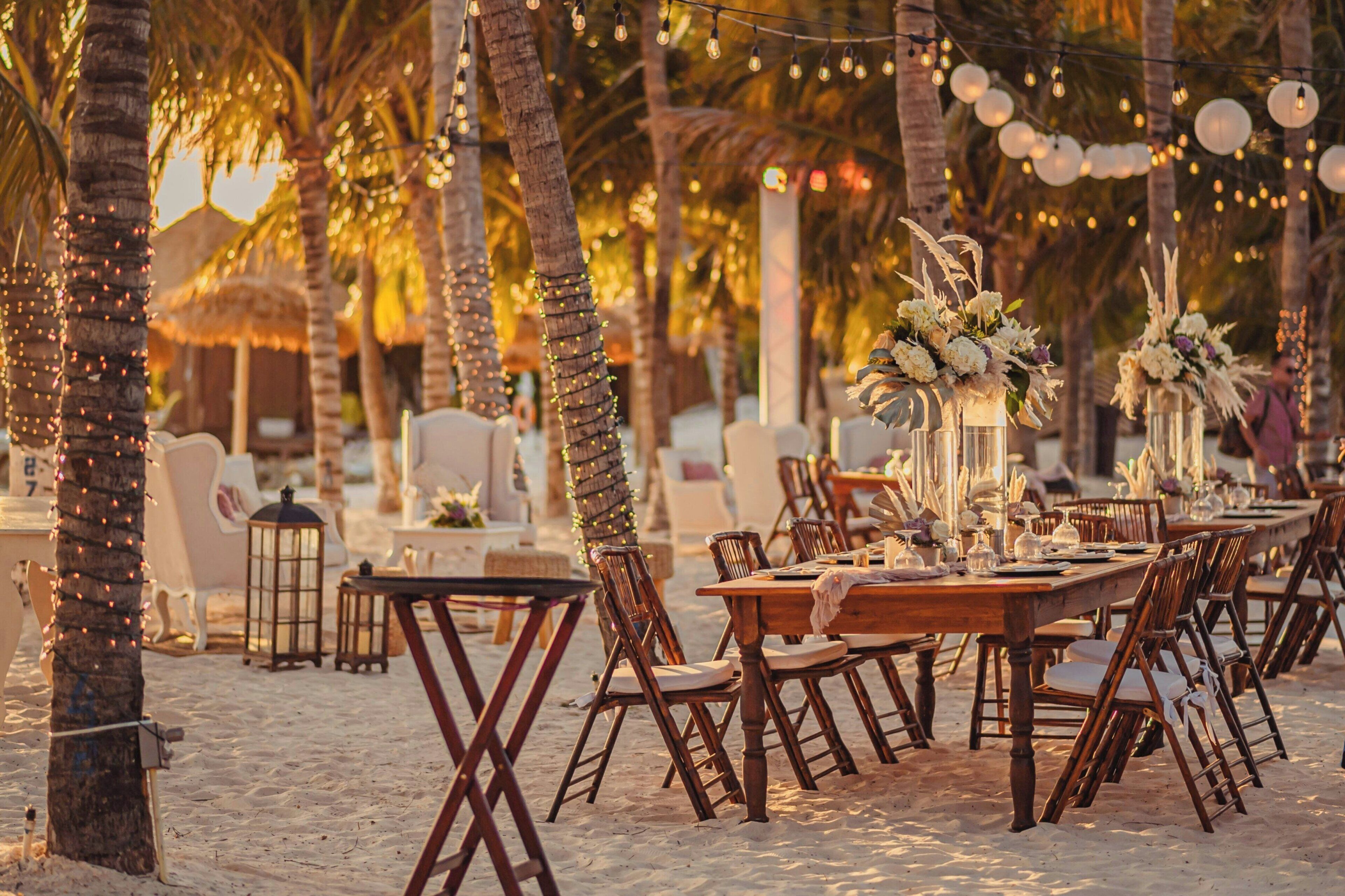 An outdoor dining area with wooden tables and chairs, surrounded by palm trees and string lights.