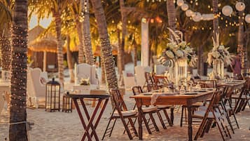 An outdoor dining area with wooden tables and chairs, surrounded by palm trees and string lights.