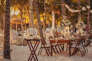 Un área de comedor al aire libre con mesas y sillas de madera, rodeada de palmeras y luces colgantes.