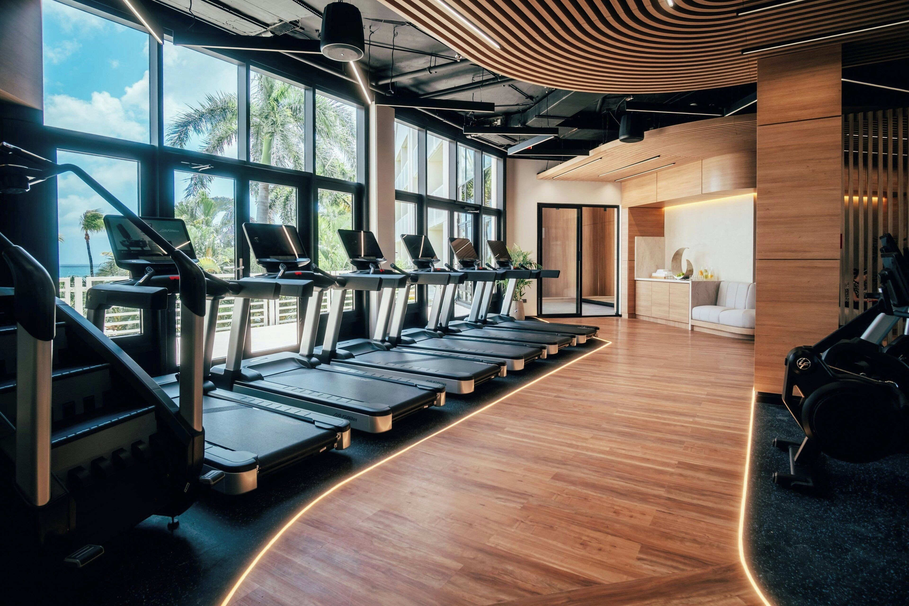 A gym with treadmills aligned along a wooden floor, large windows, and a modern ceiling design.