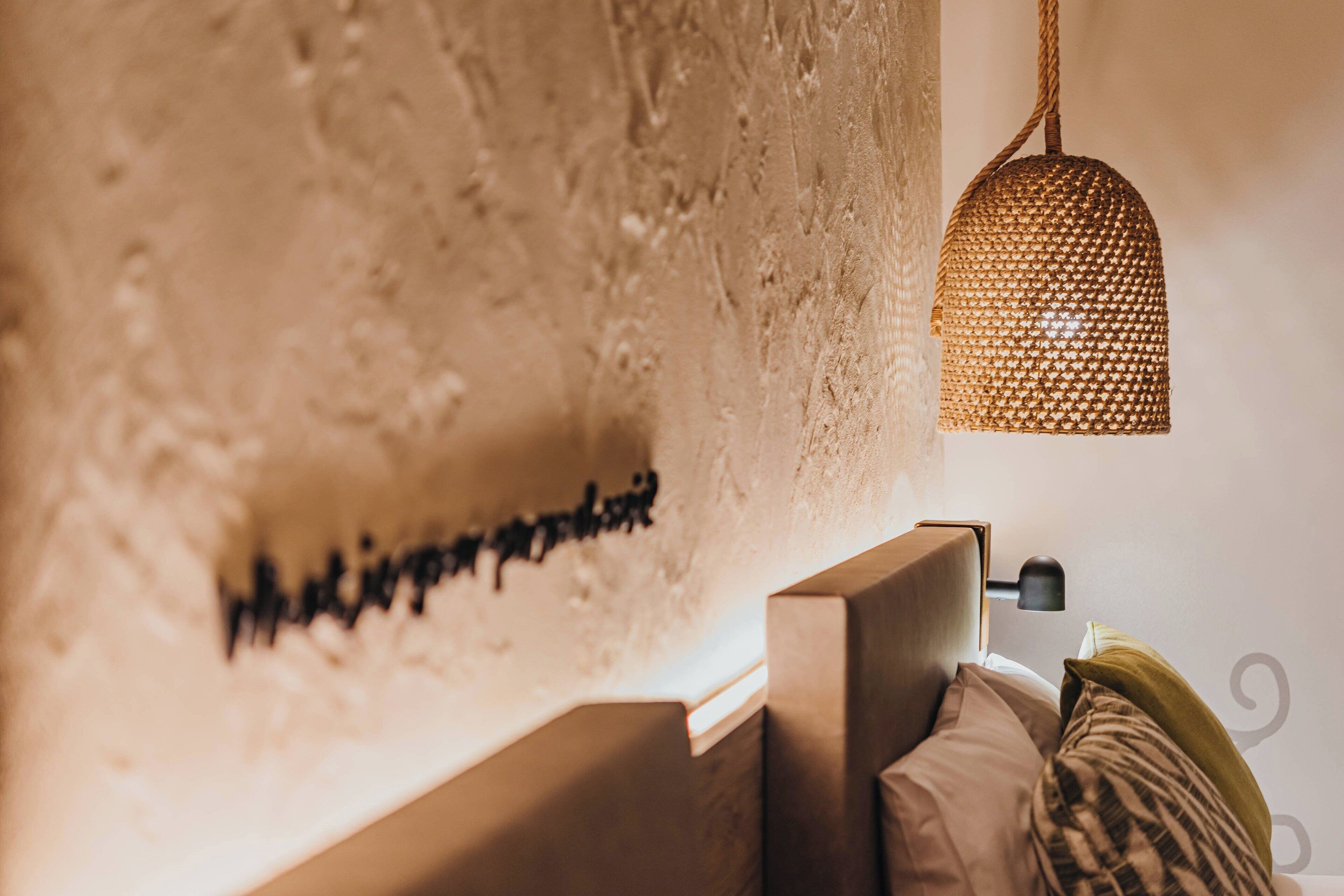 A bedroom with a textured wall, a hanging woven light fixture, and a bedside table with a lamp.