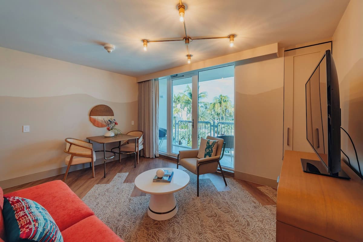 A modern living room with a sofa, coffee table, and a TV. There is a balcony with a view of palm trees.