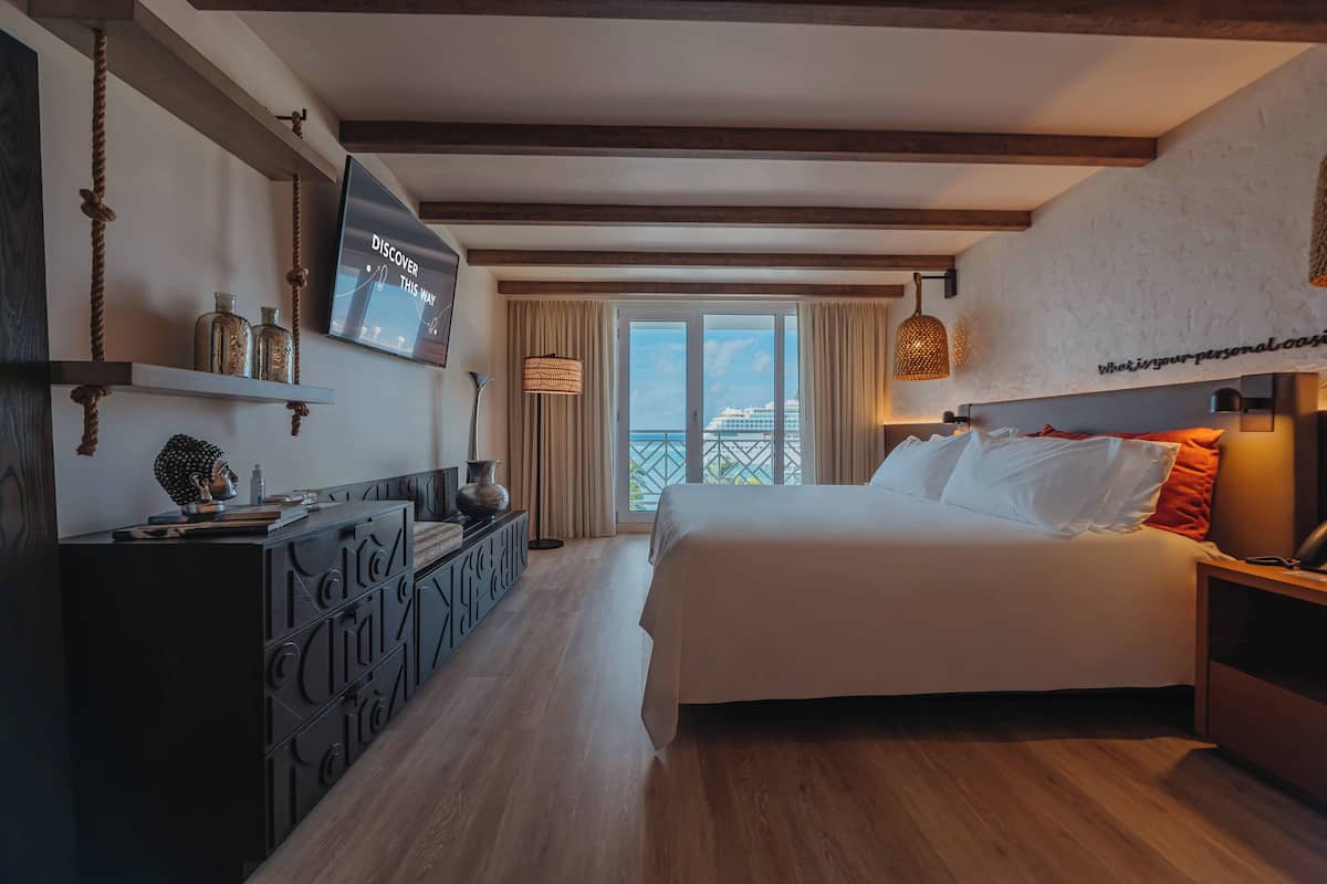 A hotel room with a bed, a TV mounted on the wall, a desk with a lamp, and a view of the sea through the window.