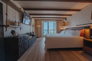 A hotel room with a bed, a TV mounted on the wall, a desk with a lamp, and a view of the sea through the window.