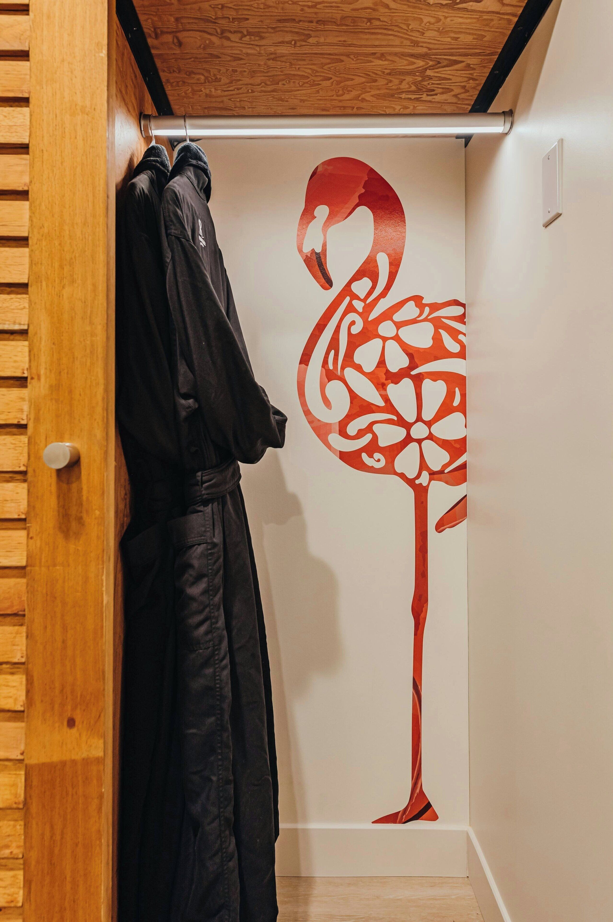 Een kast met een houten deur, een kapstok met een zwarte badjas en een muur met een muurschildering van een rode flamingo.