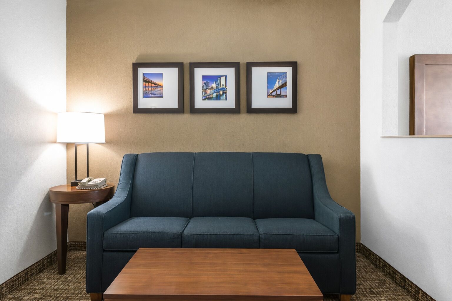 A hotel room with a blue sofa, a wooden coffee table, a lamp, and three framed pictures on the wall.