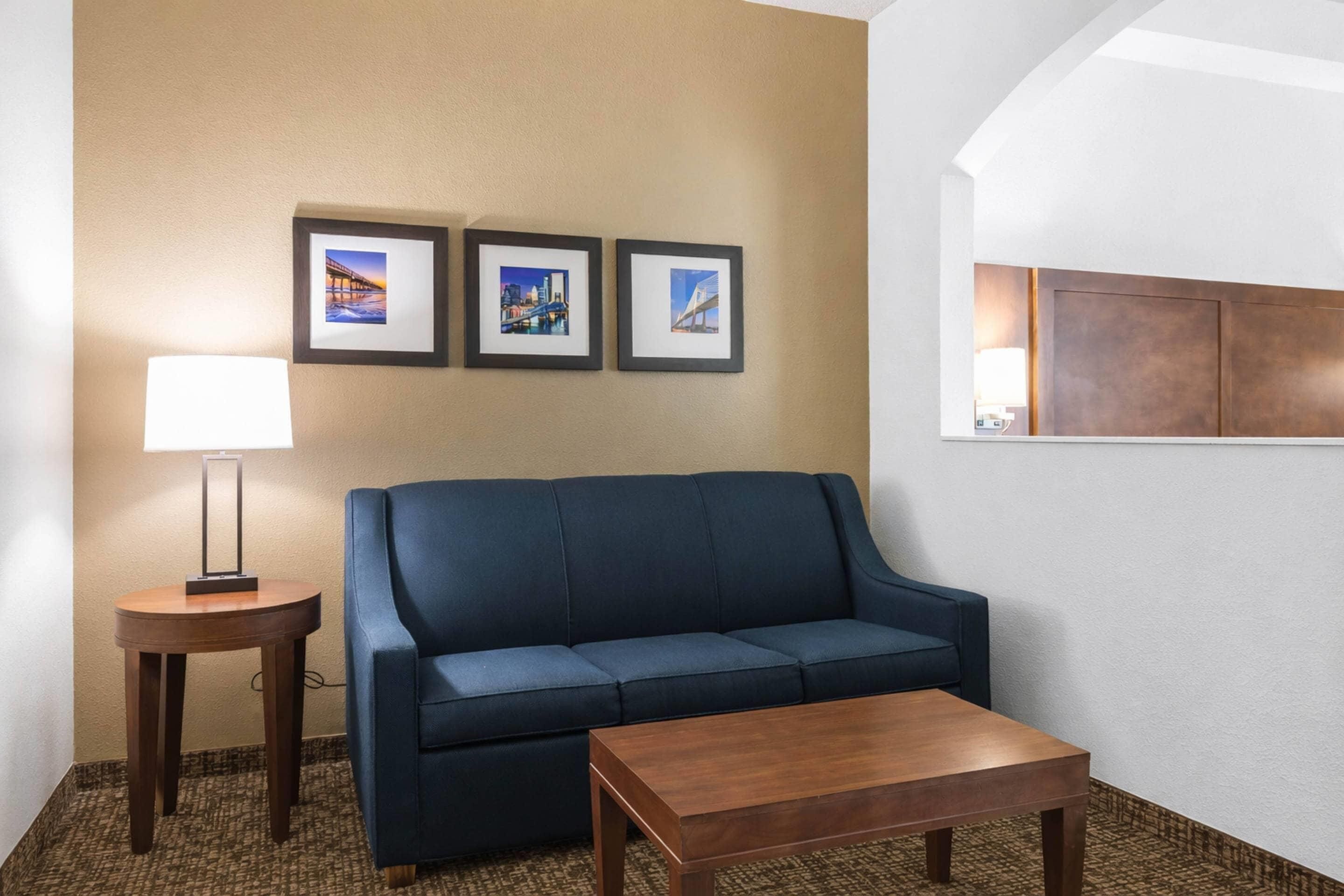 Habitación de hotel con un sofá azul, una mesa de centro de madera, una lámpara y tres cuadros en la pared.