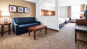 A hotel room with a blue sofa, a wooden coffee table, a bed with white linens, a TV, and framed pictures on the wall. - Comfort Suites Baymeadows Near Butler Blvd (Jacksonville)