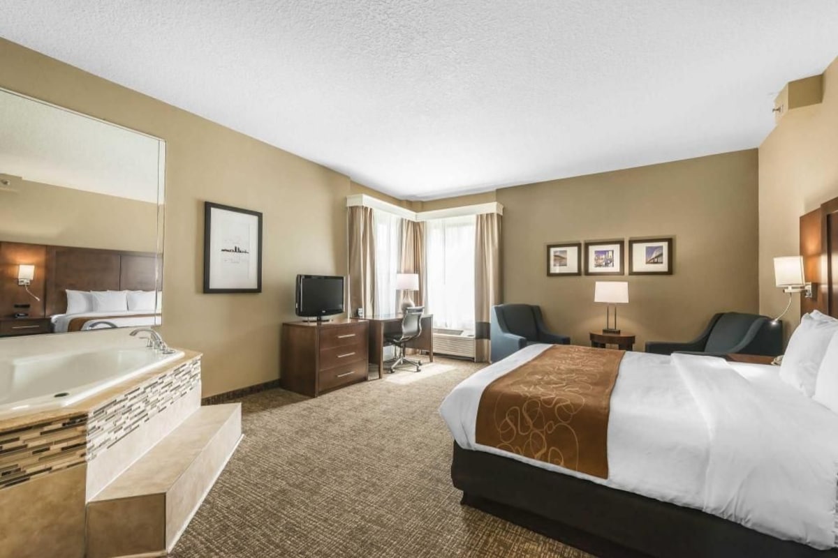 A hotel room with a large bed, a bathroom with a bathtub, a desk with a chair, a television, and framed pictures on the wall.