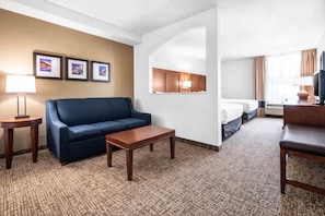 A hotel room with a blue sofa, a wooden coffee table, a bed, a desk, a lamp, and framed pictures on the wall. - Comfort Suites Baymeadows Near Butler Blvd (Jacksonville)