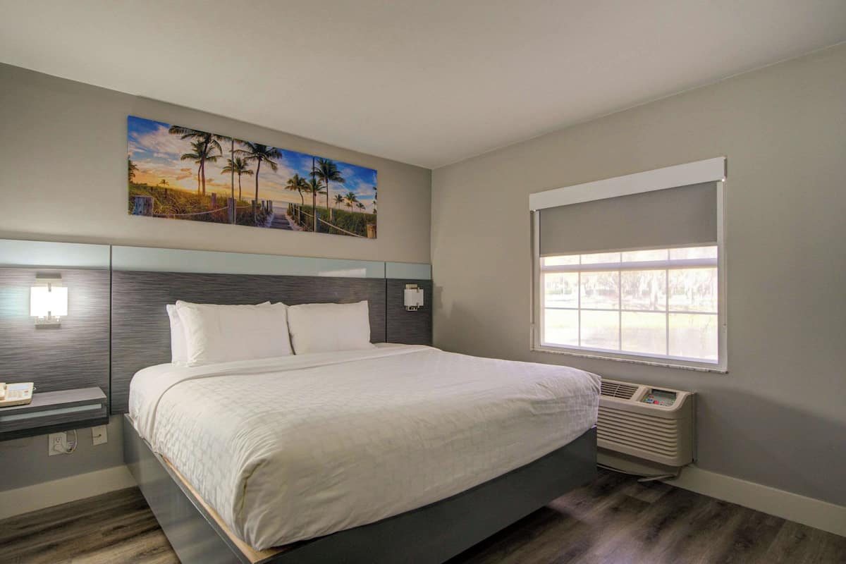 Un dormitorio con una cama grande, una mesita de noche, una lámpara de pared, un cuadro con una playa y una ventana con persianas.