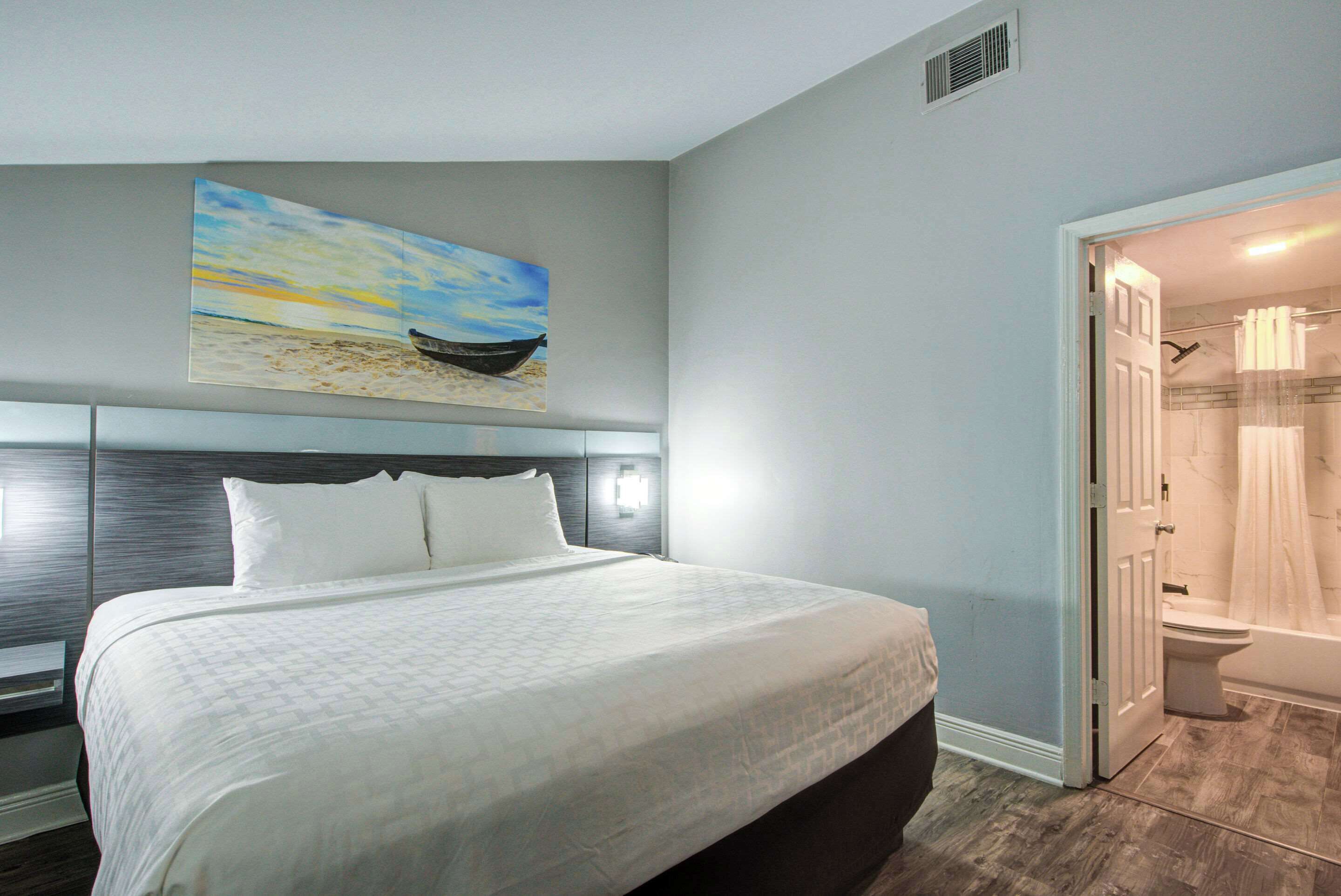 A hotel room with a large bed, a painting of a boat on the beach, a shower, and a toilet.