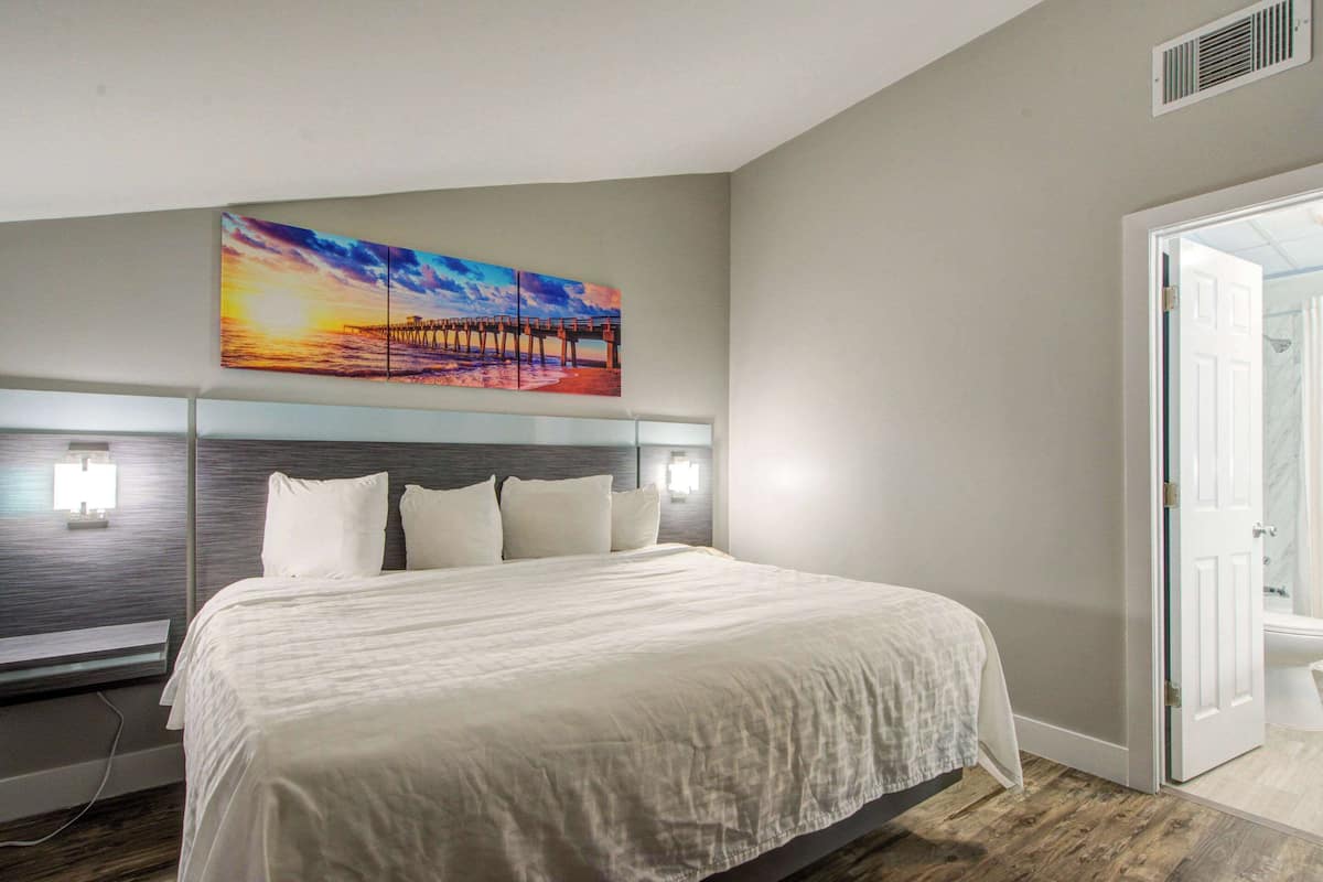 Una habitación de hotel con una cama grande, un cabecero con iluminación integrada, un cuadro de un atardecer sobre un muelle y una puerta que da al baño.