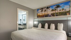 Habitació d'hotel amb un llit gran, un quadre d'una escena de platja, un capçal de llit modern i un bany visible a través d'una porta oberta.