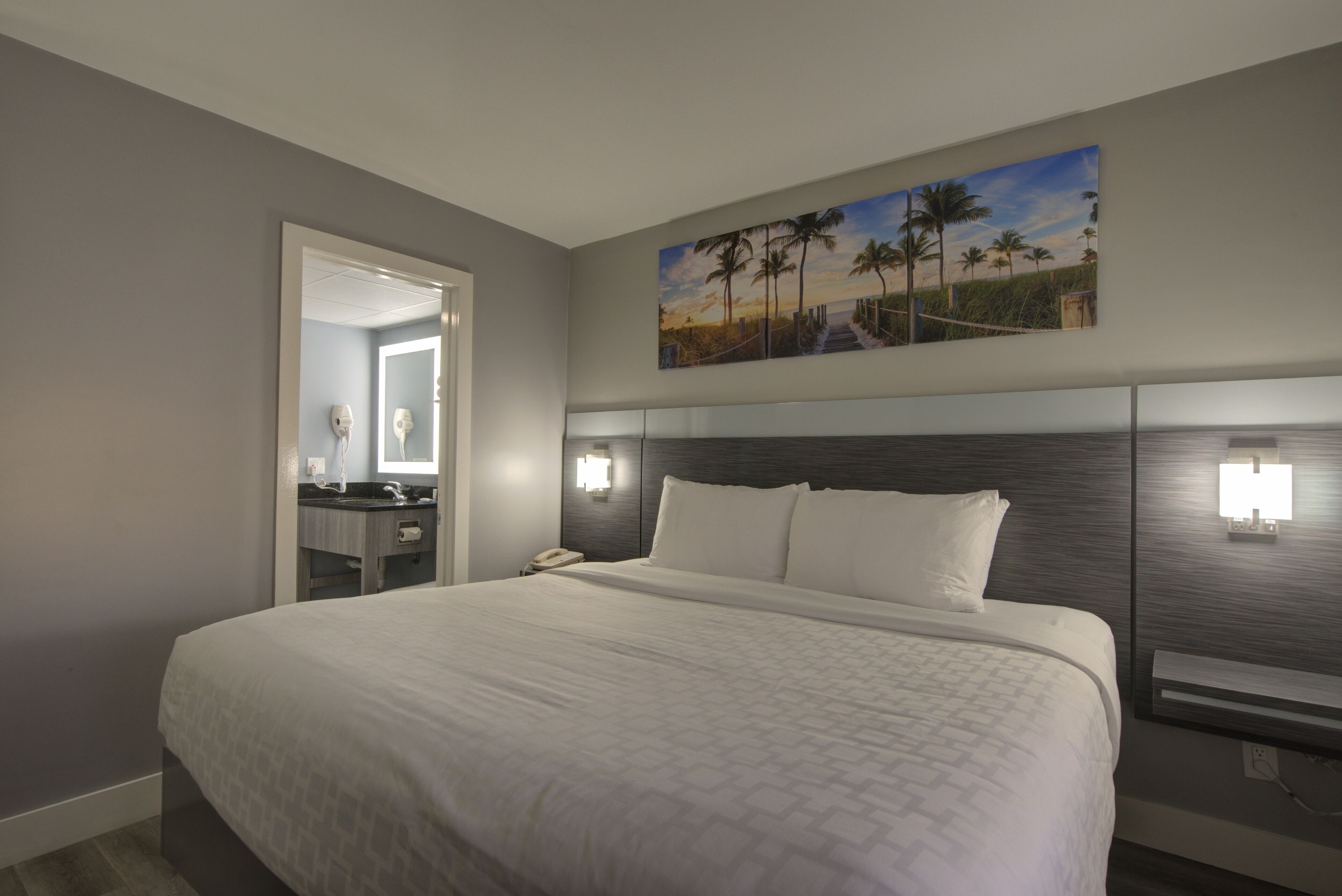 Habitación de hotel con una cama grande, una mesita de noche con lámpara, un cuadro de una playa y vista al océano a través de la ventana.
