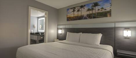 Habitación de hotel con una cama grande, una mesita de noche con lámpara, un cuadro de una playa y vista al océano a través de la ventana.