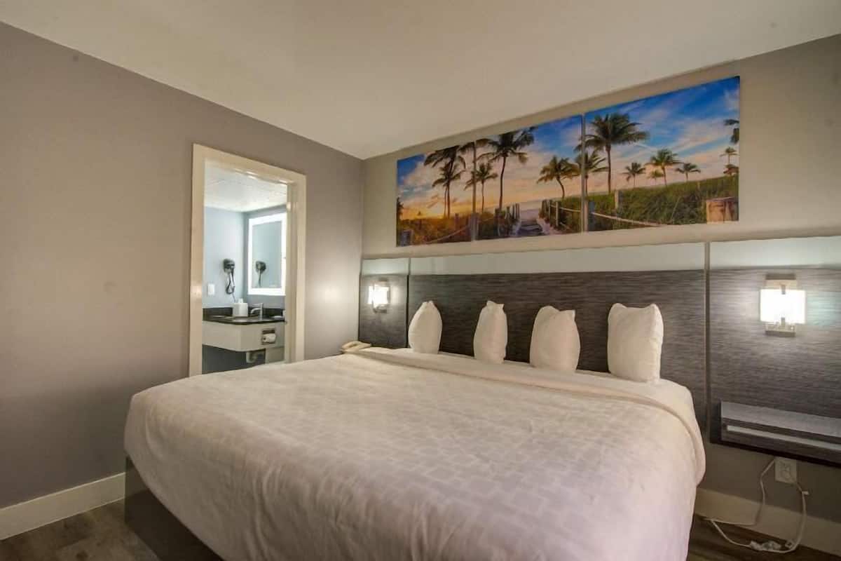 Habitación de hotel con una cama grande, un cuadro de una playa, un espejo y una mesita de noche con una lámpara.