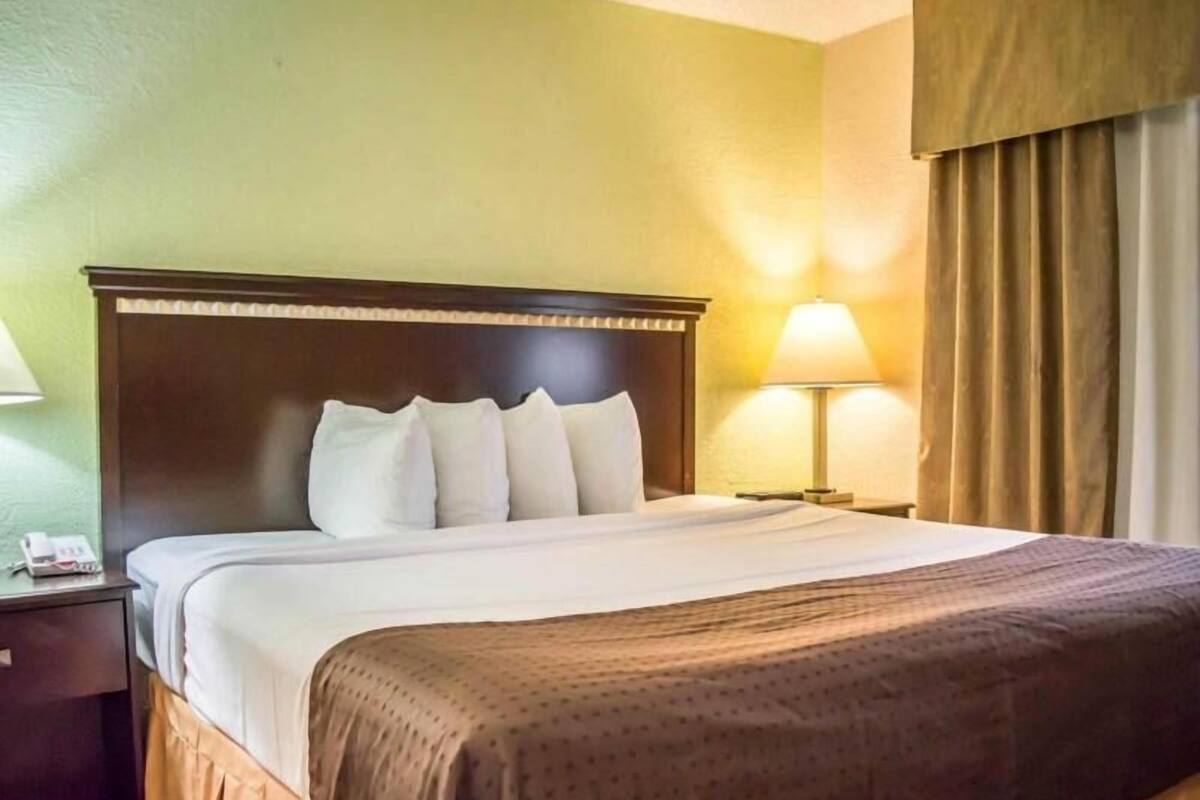Habitación de hotel con cama, mesita de noche, lámpara y ventana con cortinas.