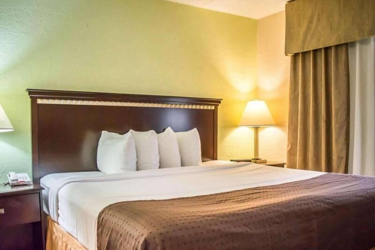 Una habitación de hotel con una cama, una mesita de noche, una lámpara y una ventana con cortinas.
