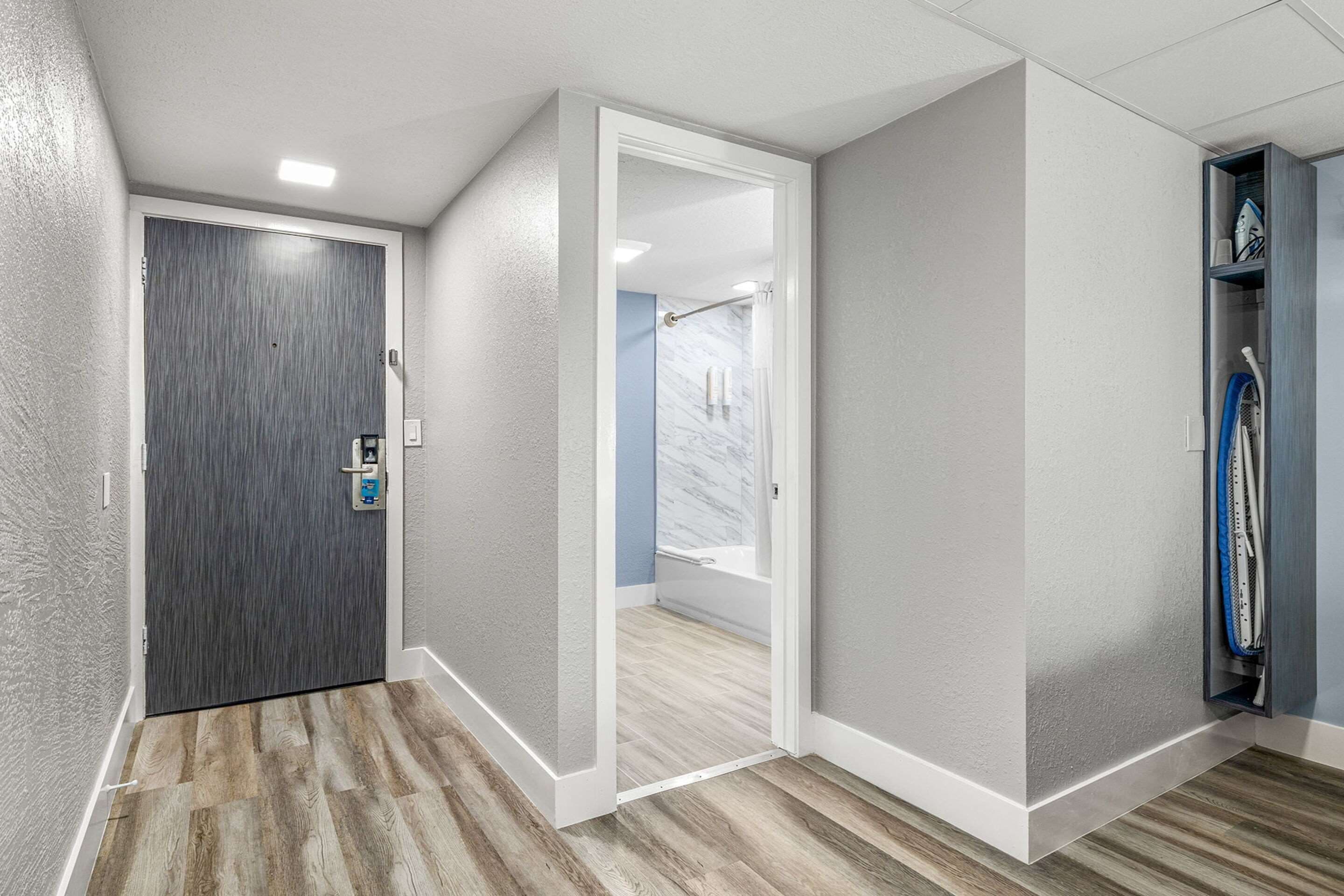 A hallway with a wooden floor, a grey door, and a white-framed door leading to a bathroom.
