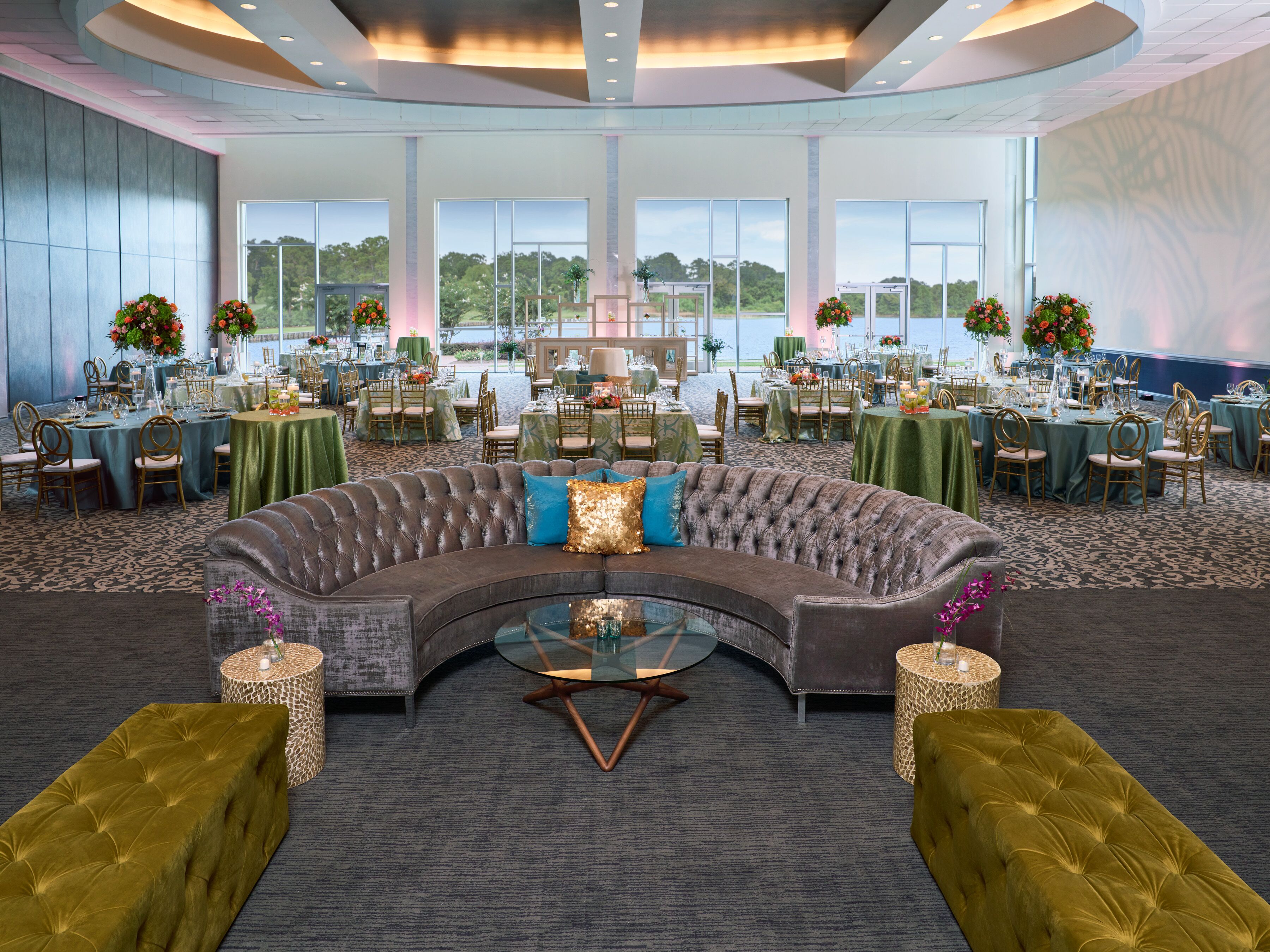 Une salle de banquet spacieuse et élégamment aménagée, avec un canapé d’angle incurvé, des tables rondes et des chaises, le tout mis en valeur par de grandes fenêtres offrant une vue panoramique.