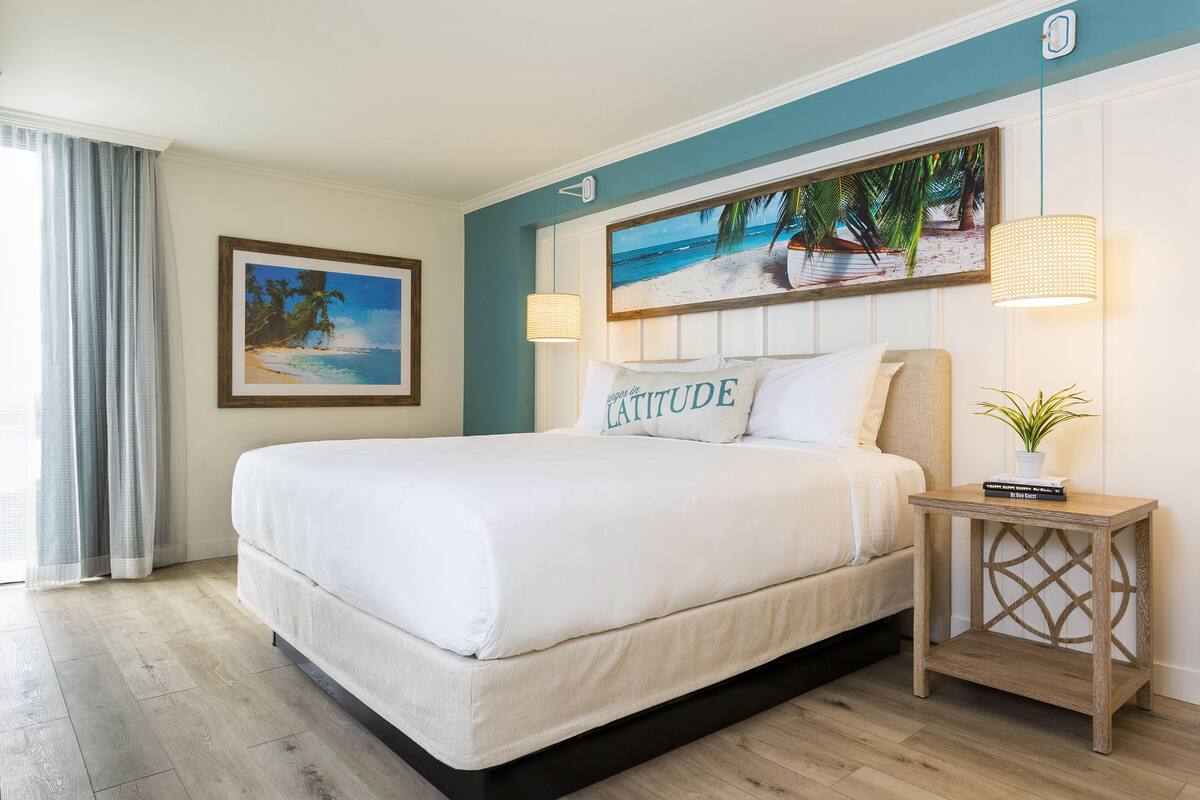 A neatly made bed with a pillow labeled "LA TATUDE" in a room with a wooden floor, a painting of a beach scene, and a small table with a plant.
