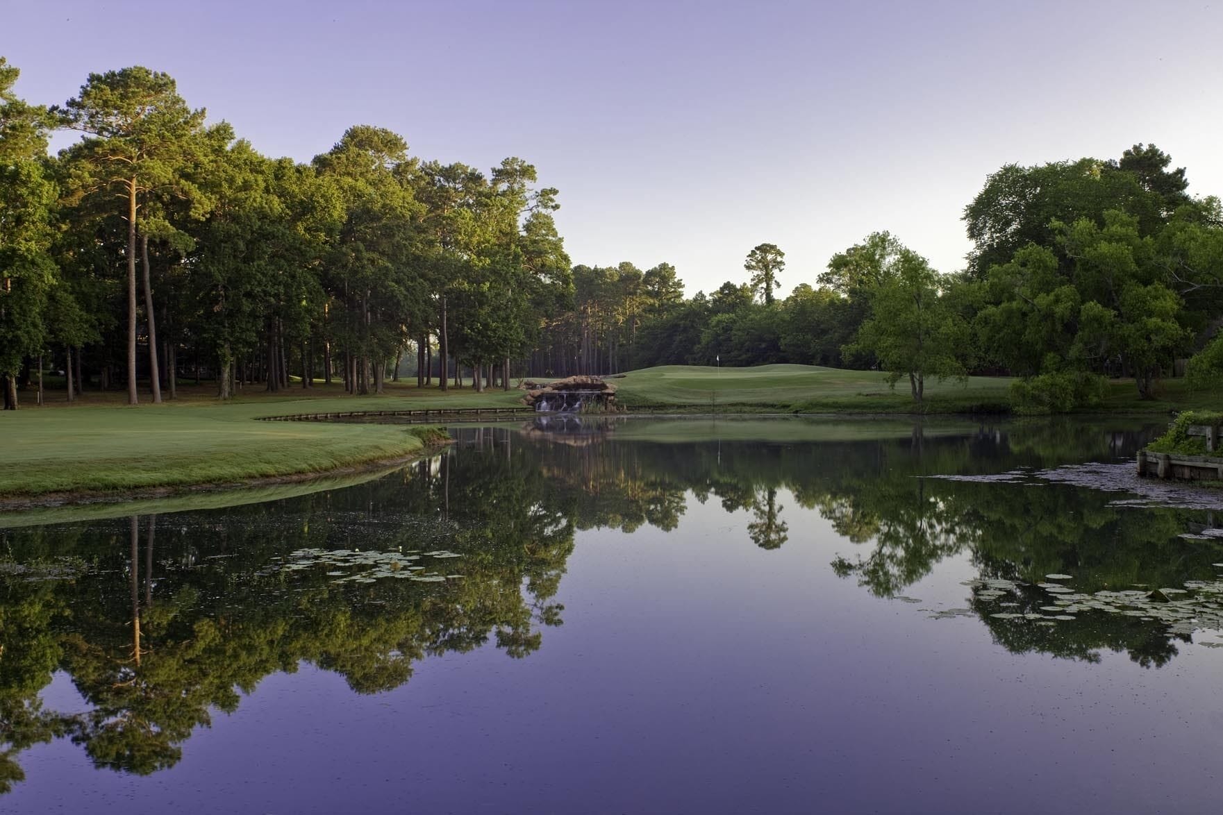 A serene lakeside view with a golf course, calm water, and lush greenery.