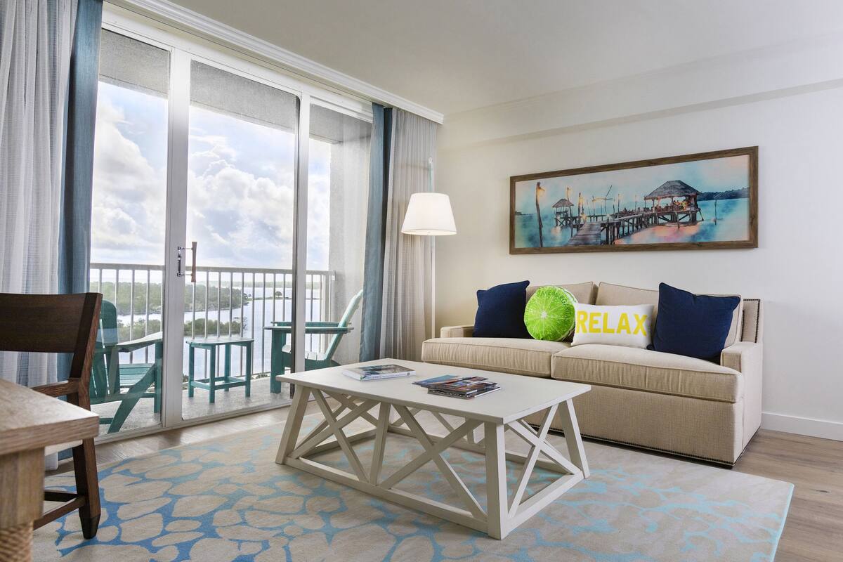 A room with a sofa, a coffee table, a painting, and a view of the outdoors.