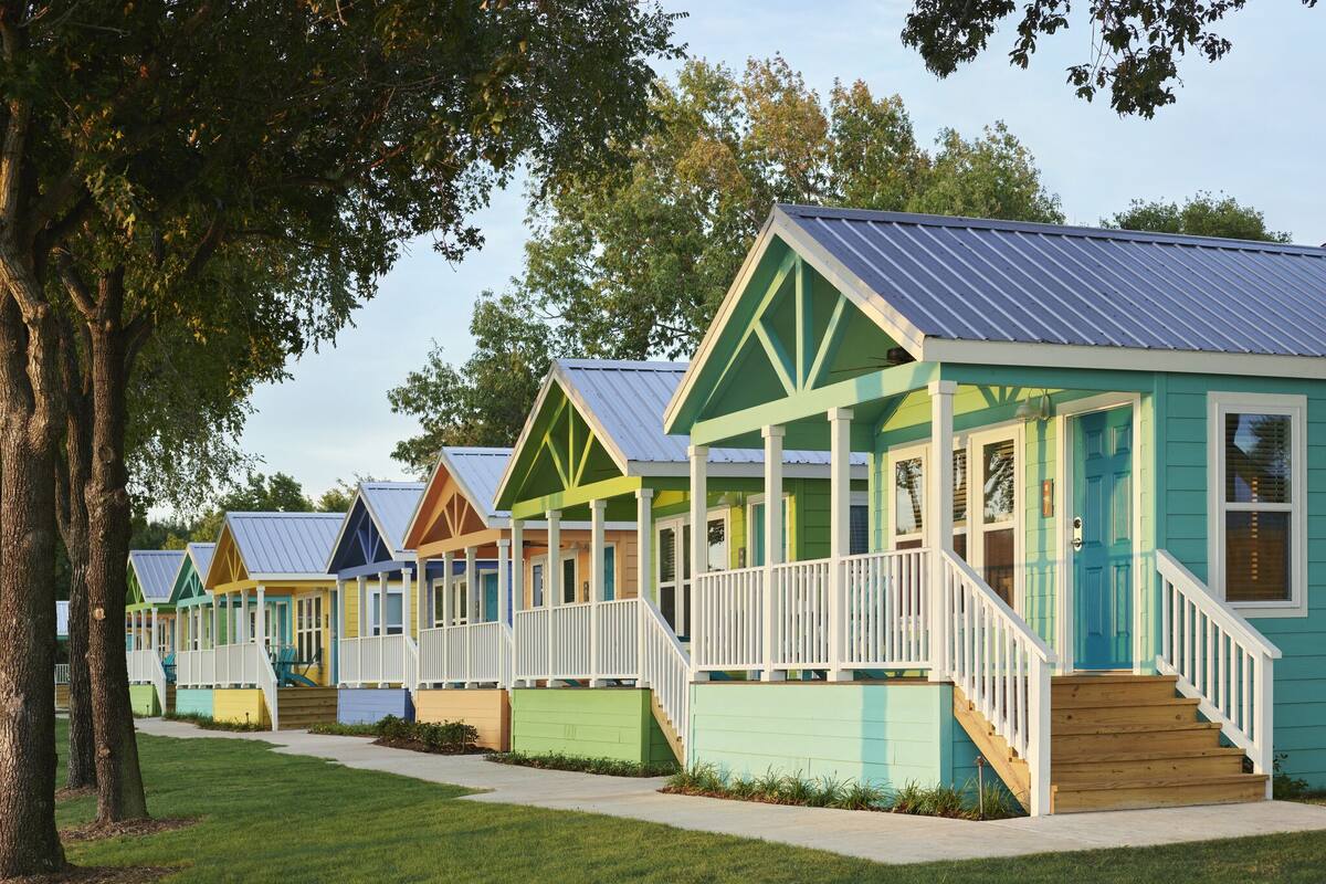 A row of colorful cabins with white railings and a covered porch, set in a grassy area with trees.