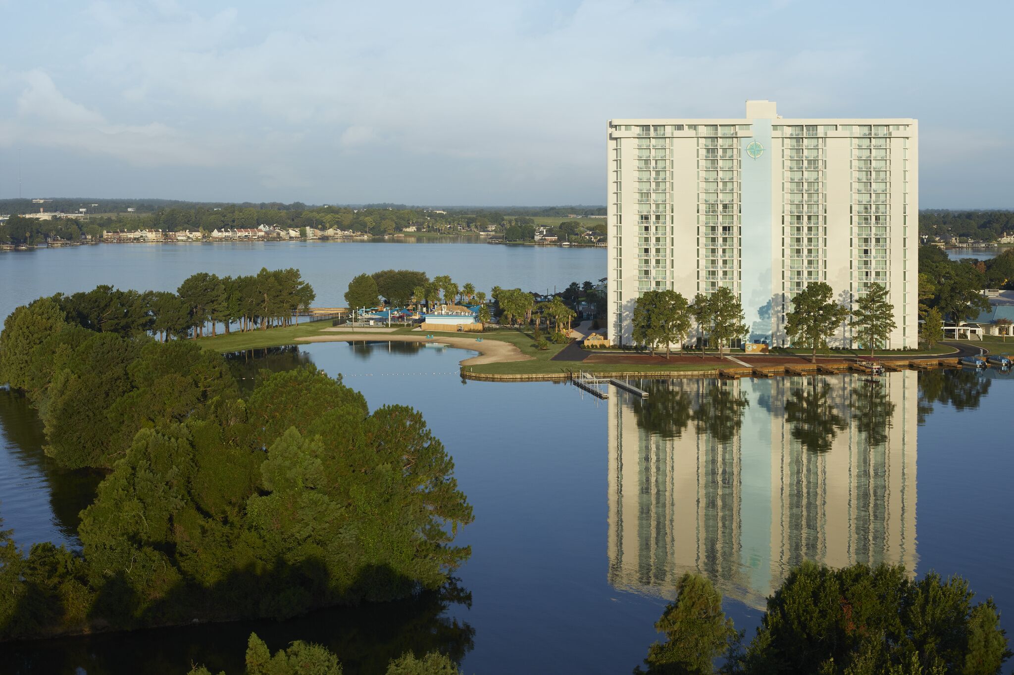 A high-rise hotel with a reflection in the water, surrounded by trees and a small island with a dock.
