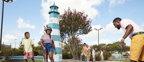 A group of people playing croquet on a blue field with a lighthouse and trees in the background.