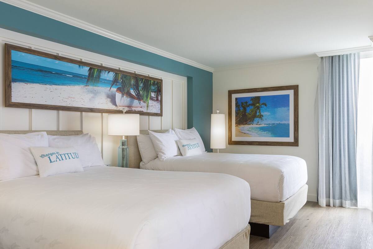 A hotel room with two beds, a large beach-themed painting above the beds, and a smaller painting on the wall.
