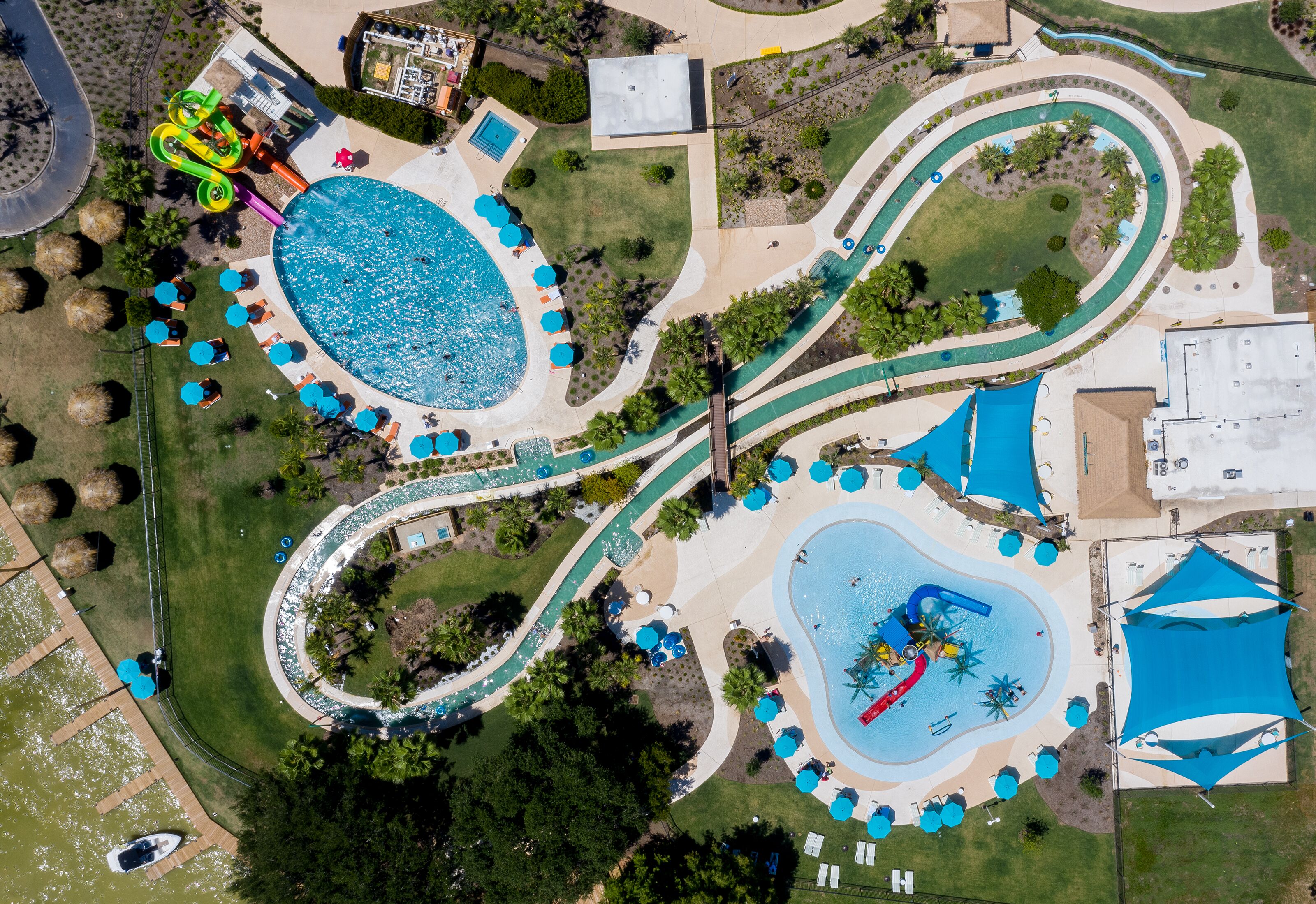 Vista aérea de un parque acuático con múltiples piscinas, senderos sinuosos y zonas de sombra con sombrillas.