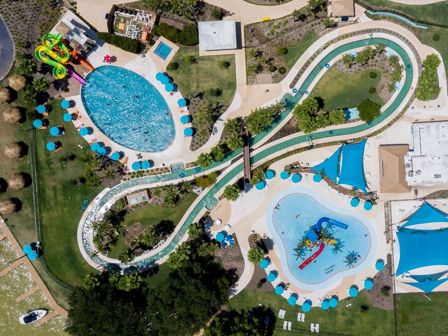 Vista aérea de un parque acuático con múltiples piscinas, senderos sinuosos y zonas de sombra con sombrillas.