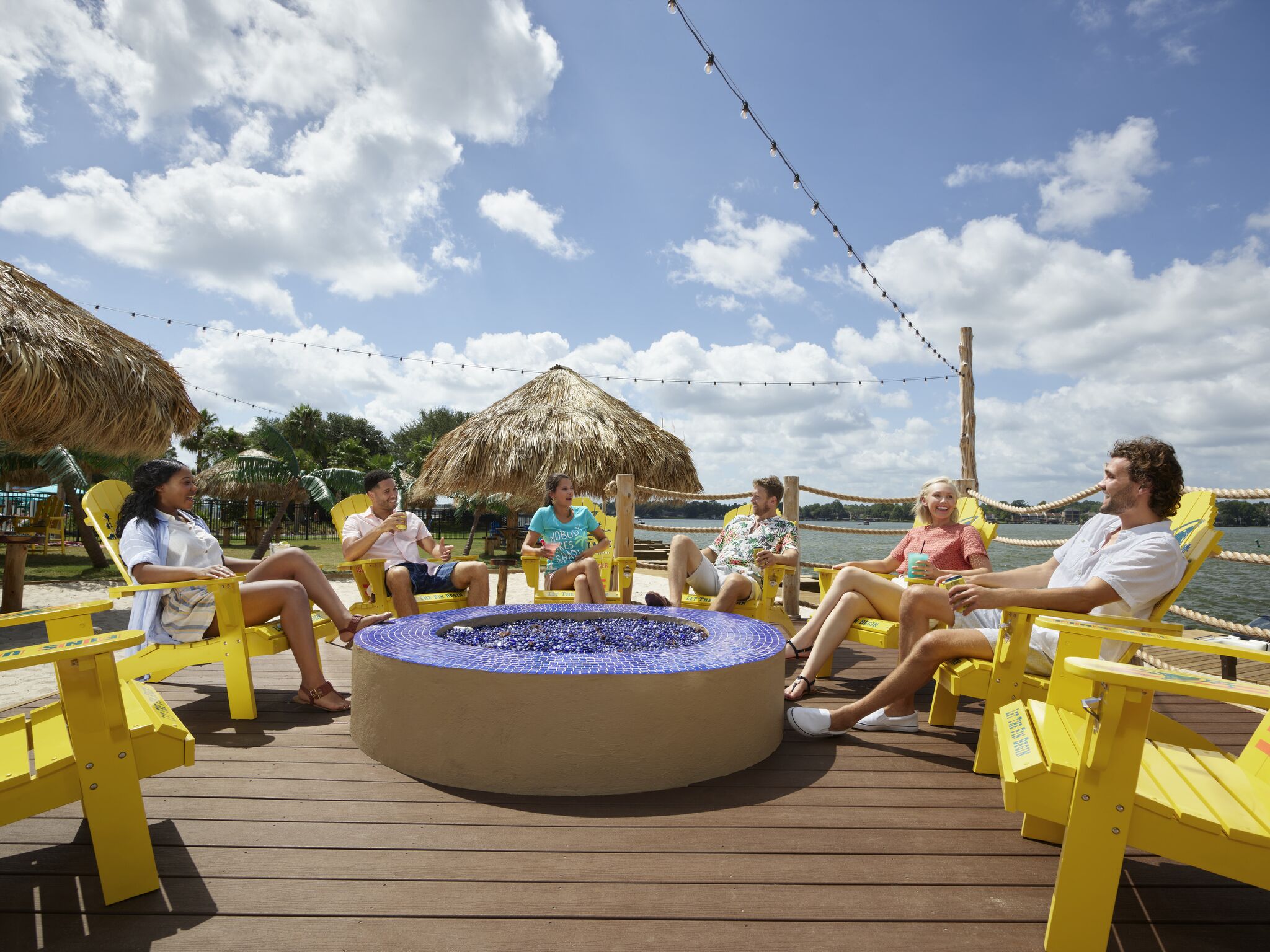 A group of people sitting around a circular fire pit on a wooden deck with thatched umbrellas in the background.