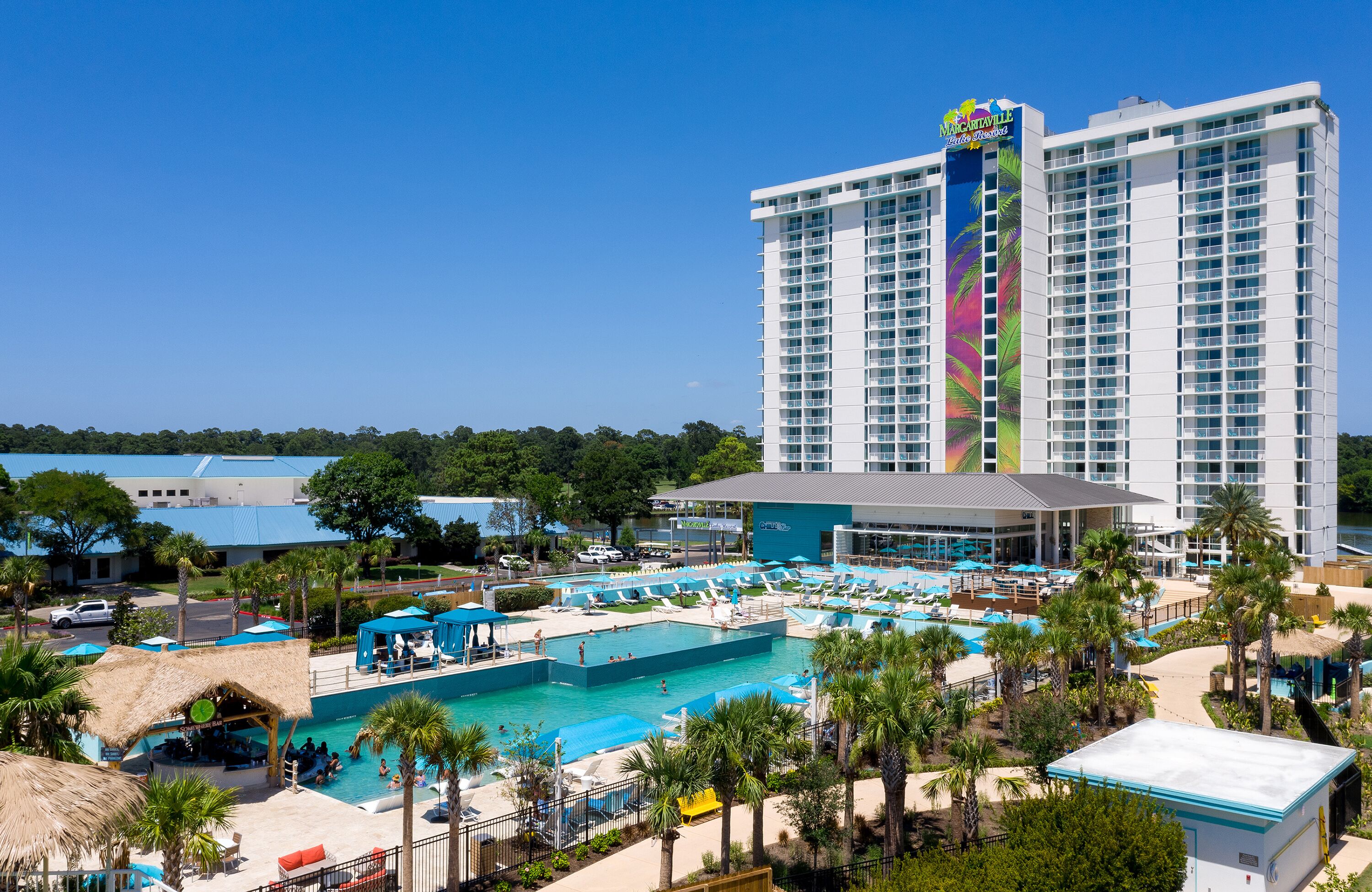 A large hotel with multiple swimming pools, a beach area, and a tall building with a colorful sign.
