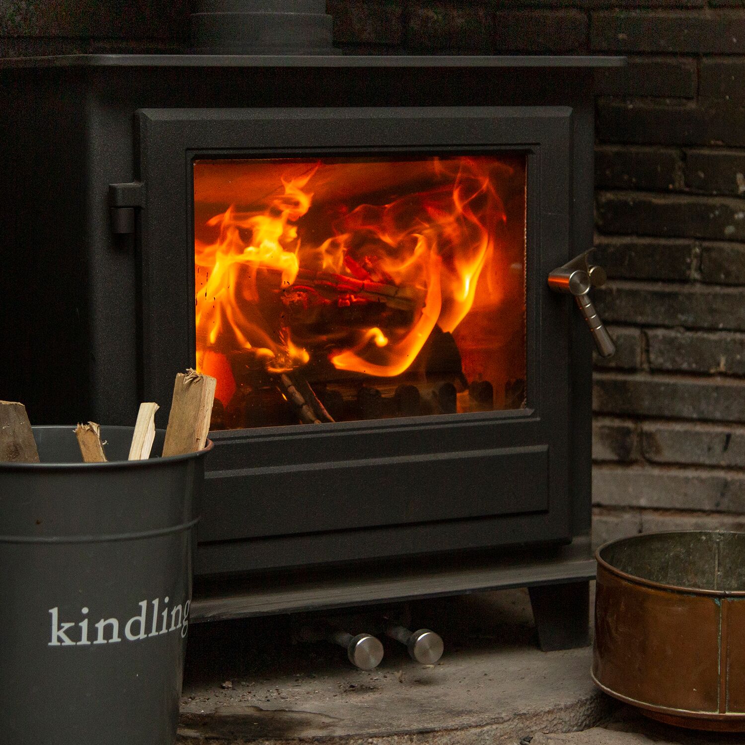 A wood-burning stove with visible flames, a metal bucket labeled "kindling," and a metal pot on the ground.