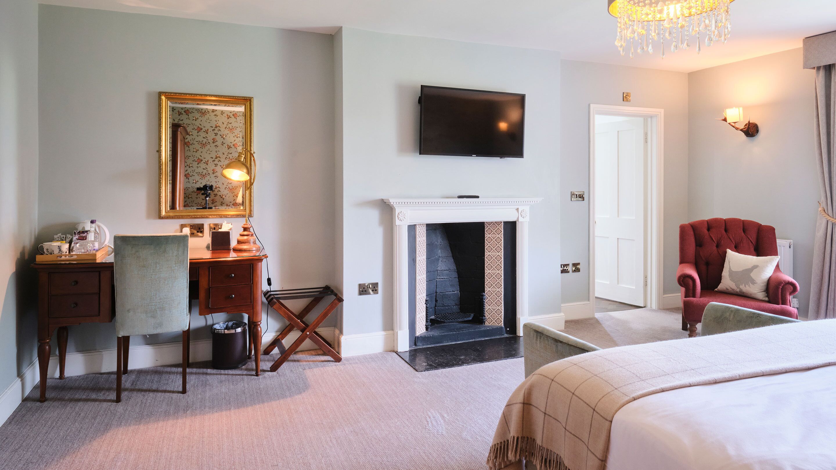 Habitación de hotel con una cama, un escritorio con silla, una chimenea, un televisor y un sillón rojo.