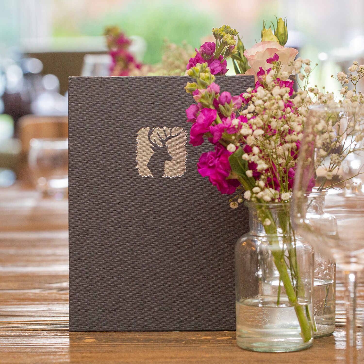 Un livre de couleur brun foncé, orné sur sa couverture de la silhouette d’un cerf, est posé sur une table en bois, à côté d’un vase en verre transparent contenant des fleurs roses et blanches.