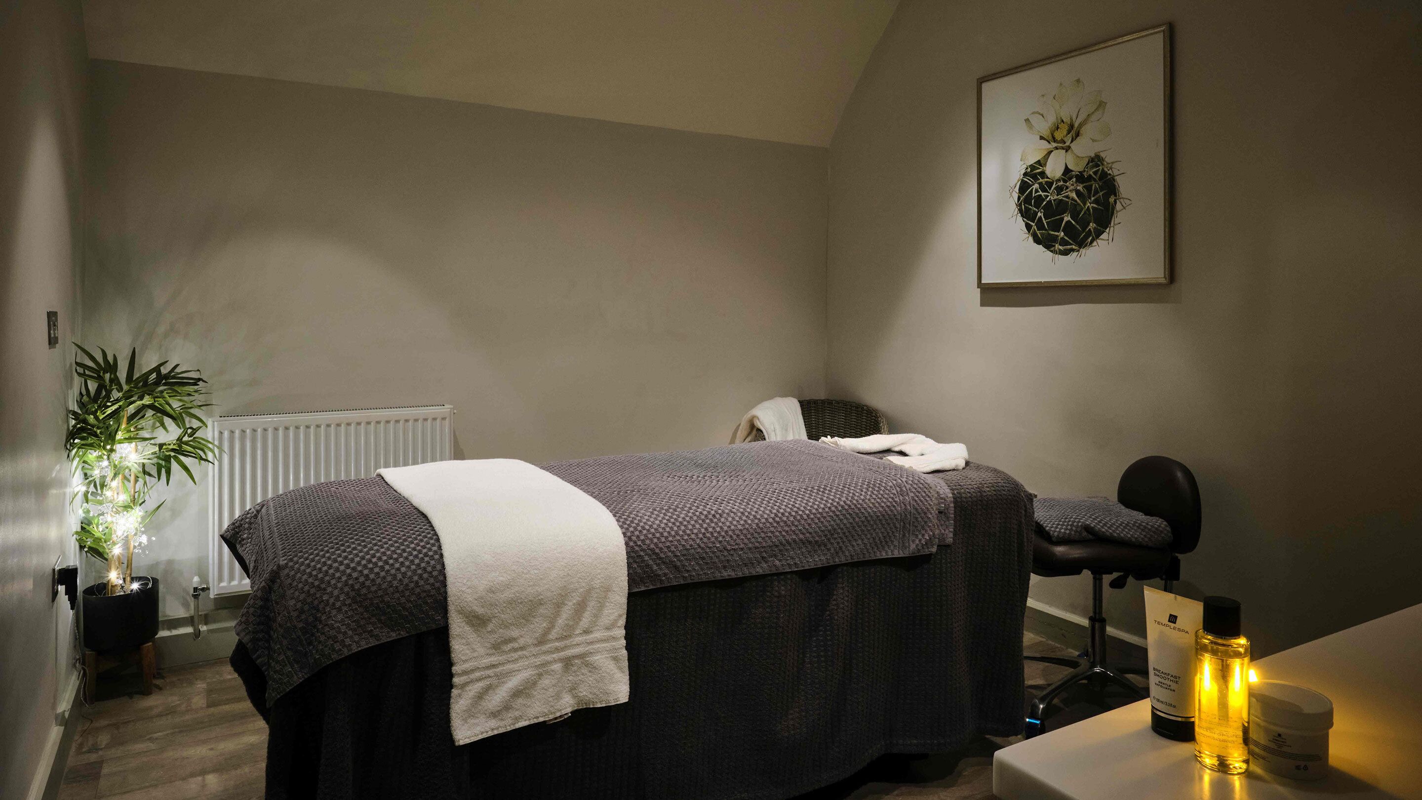 A spa room with a massage table, a chair, a potted plant, a radiator, and a framed artwork on the wall.