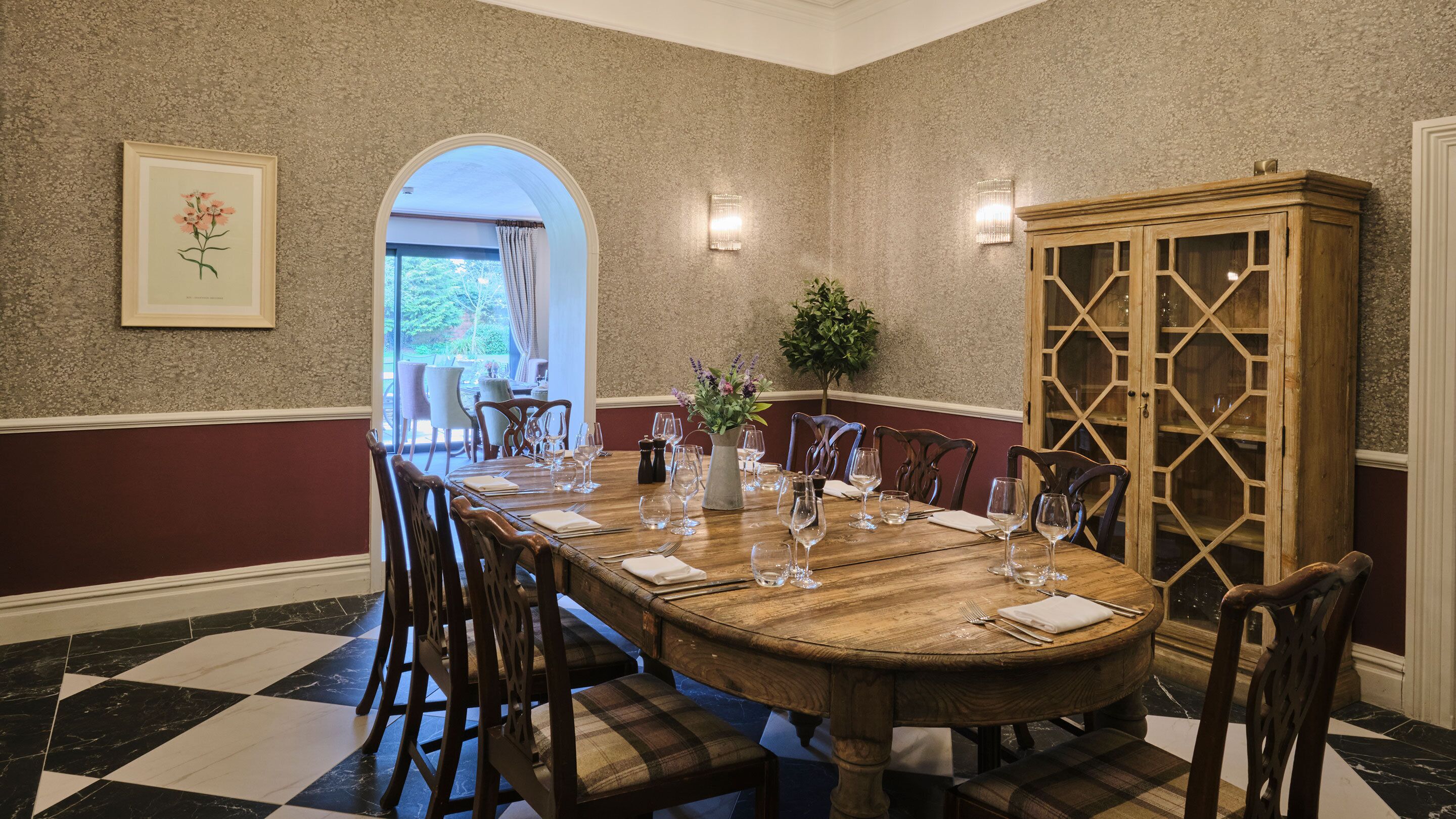 A dining room with a wooden table set for six, surrounded by chairs, a vase with flowers, and a cabinet with glass doors.