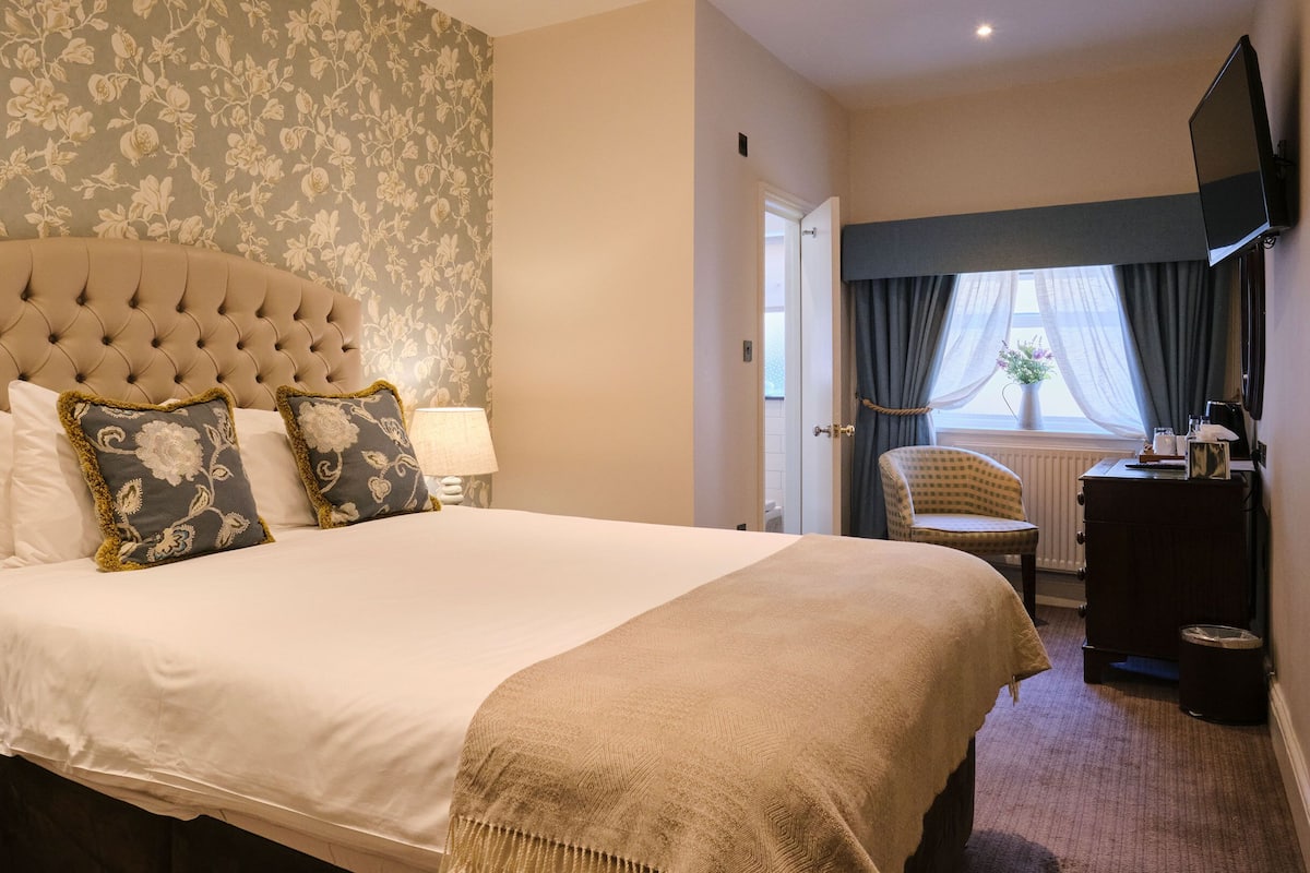 Quarto de hotel com uma cama grande, cabeceira estofada, papel de parede floral, uma janela com cortinas, uma cadeira, uma escrivaninha e uma televisão.