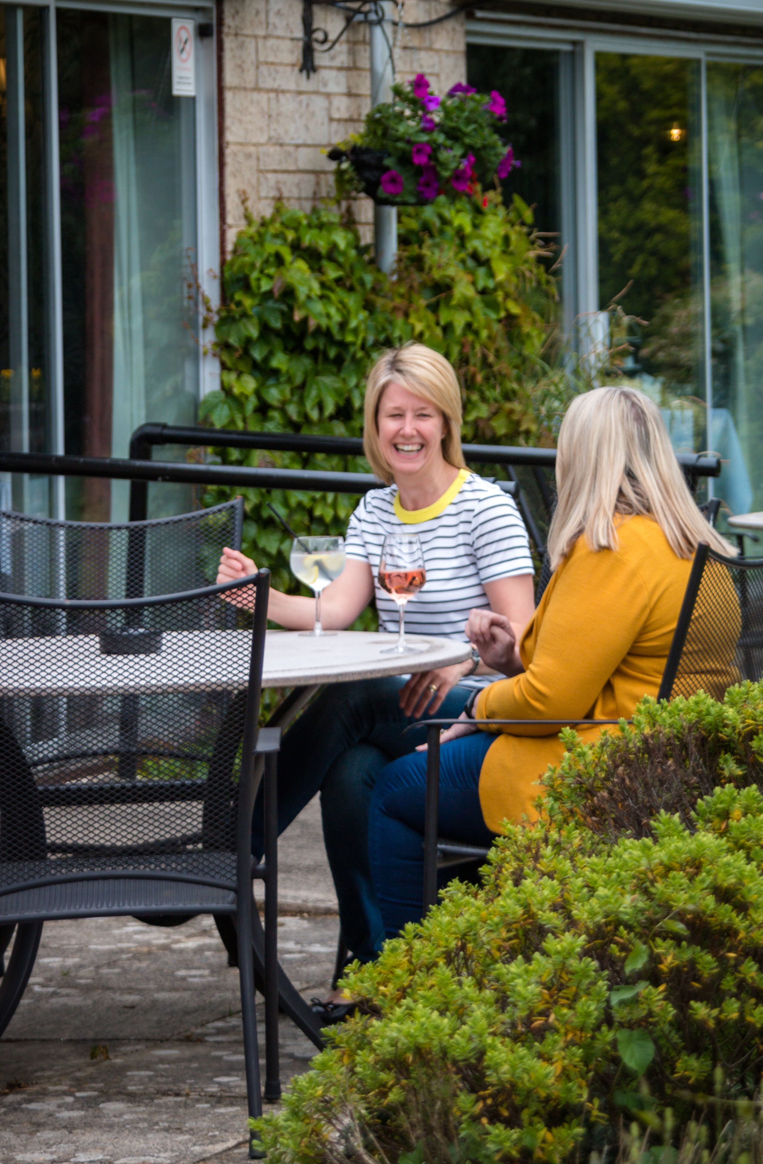 Two people seated at an outdoor table with chairs, a glass of wine, and a potted plant in the background.