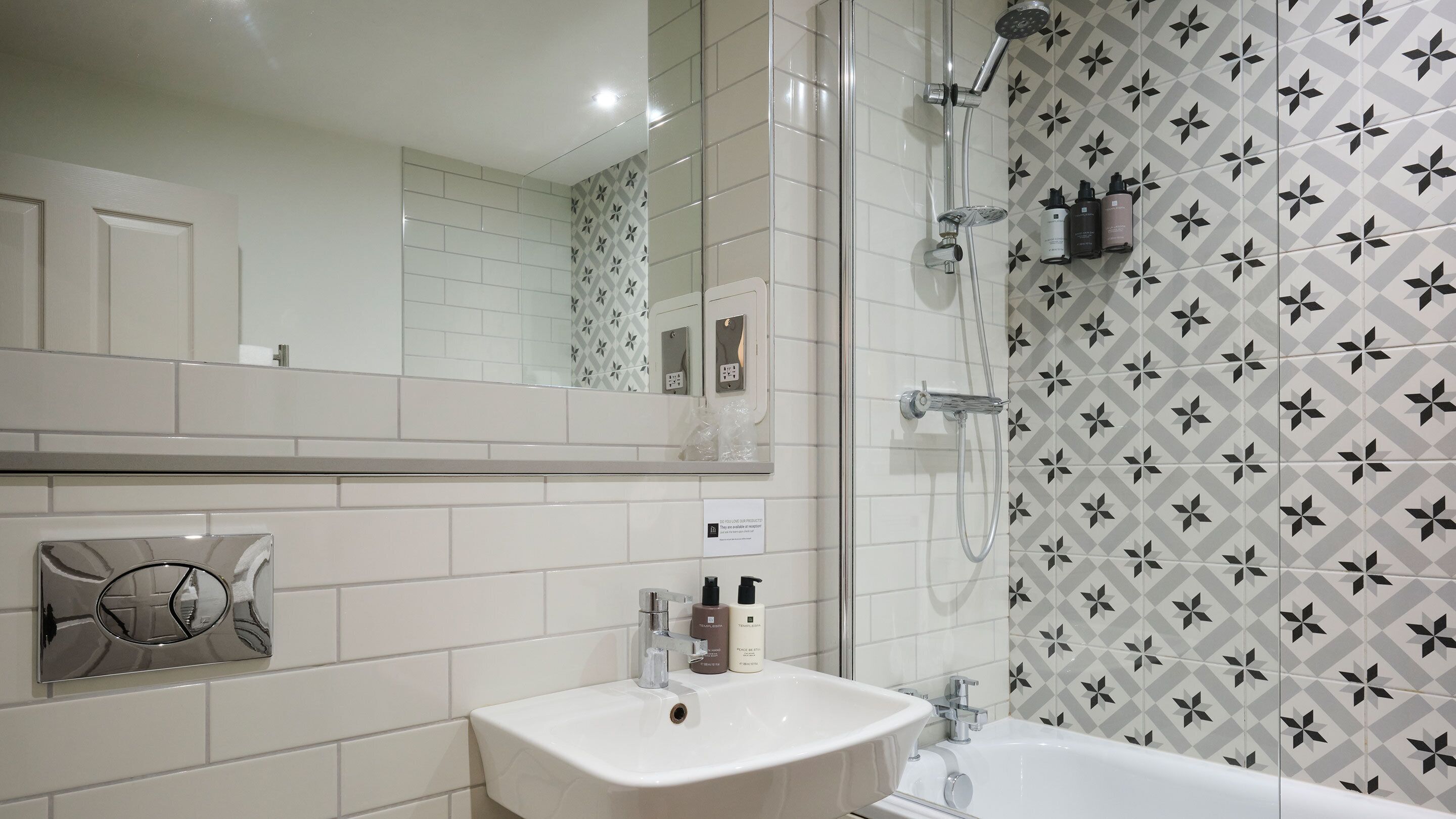 A bathroom with a white sink, a mirror, a shower, and patterned tiles.