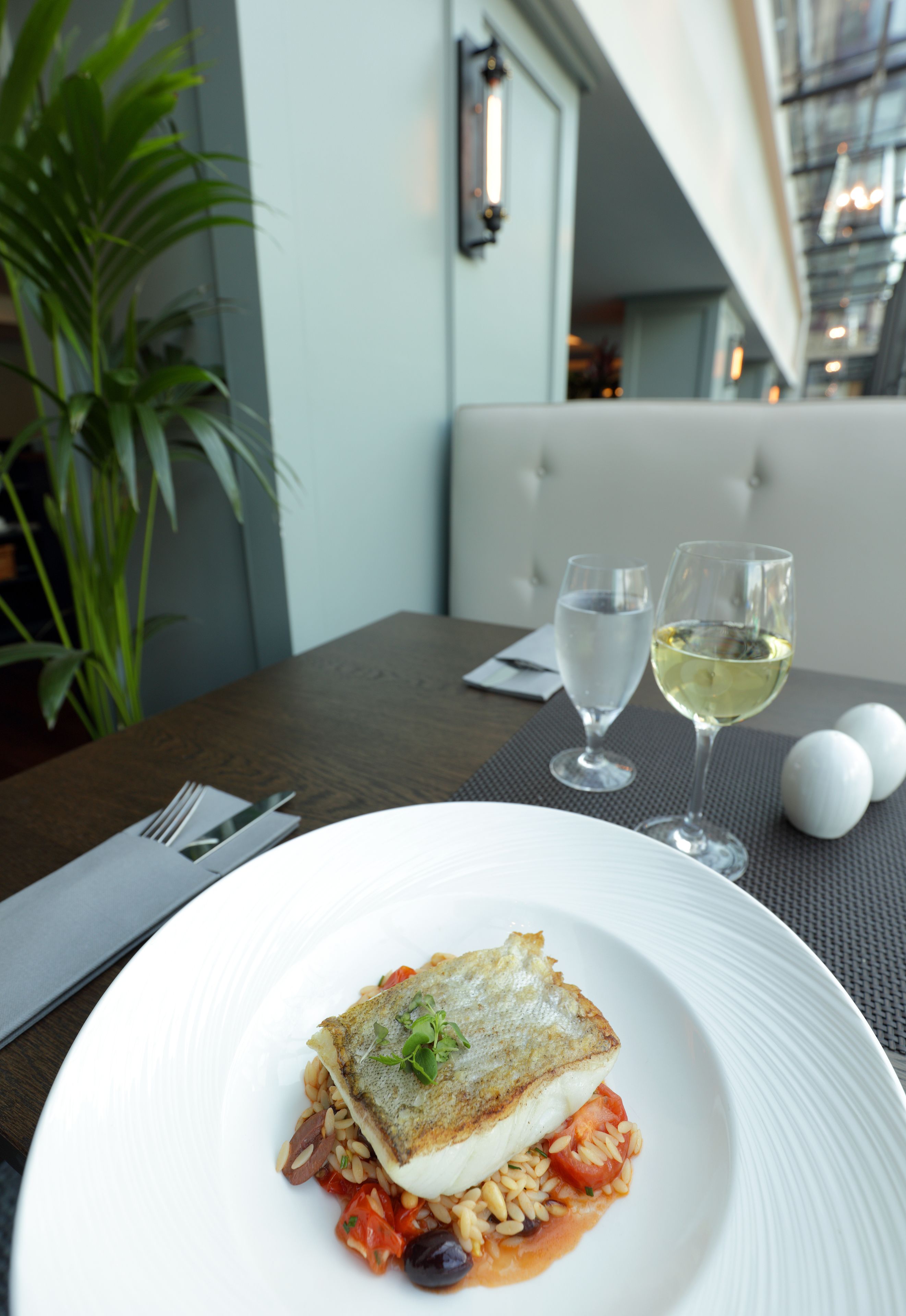 A dining table with a plate of food, two glasses of white wine, and a fork and knife set.